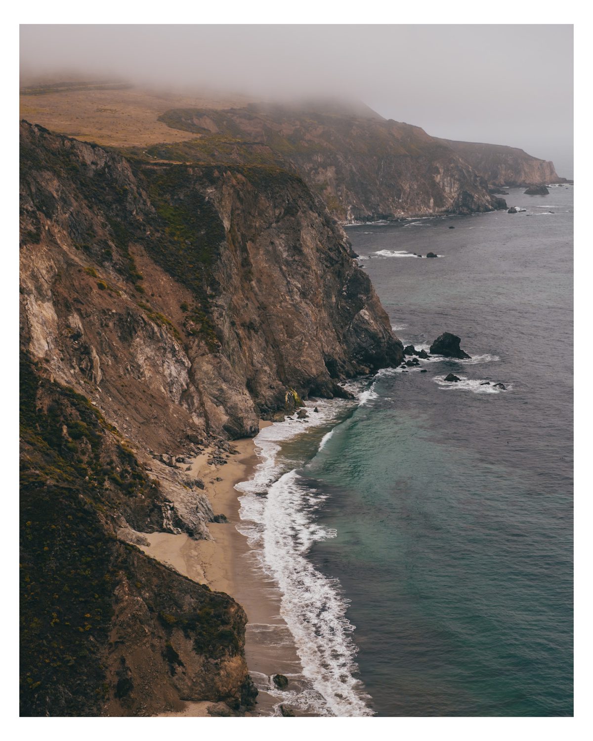 A fog-covered coastal cliff dropping to a narrow curved beach, with pale surf breaking over turquoise water.